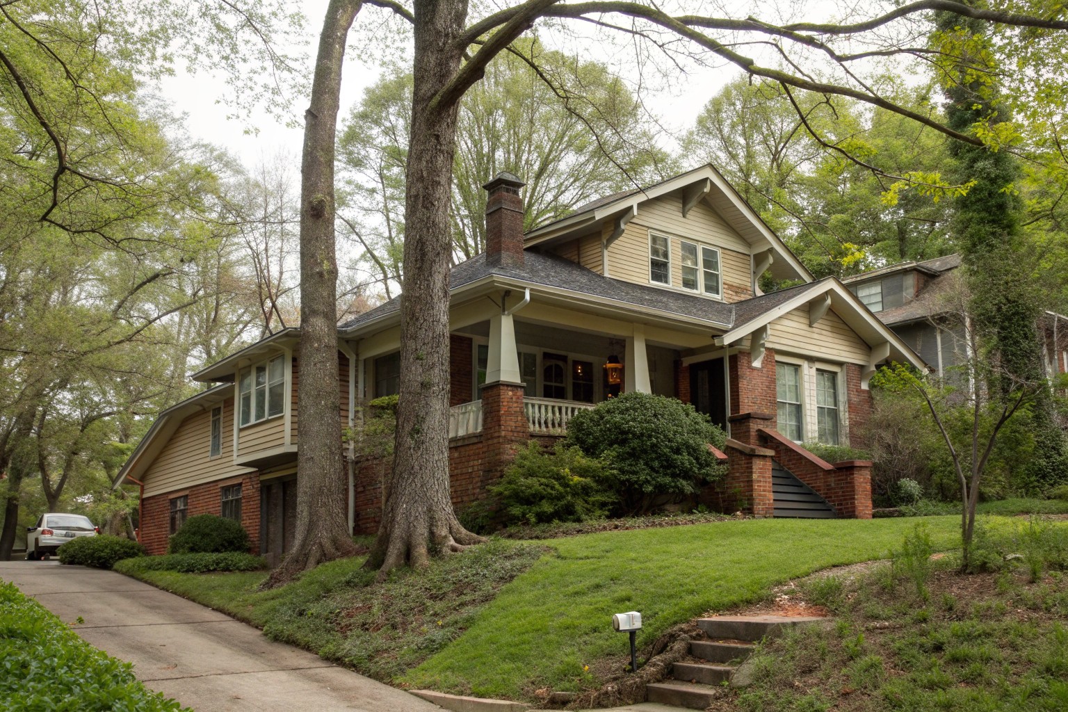 A 1920s Craftsman bungalow on a sloping lot with a brick daylight basement under tall oaks and dogwoods